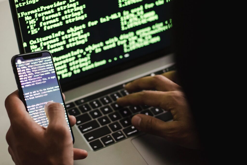 Free Unrecognizable hacker with smartphone typing on laptop at desk Stock Photo